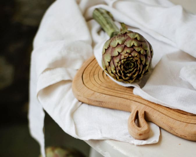 An artichoke rests on a wooden cutting board and white kitchen tea towel